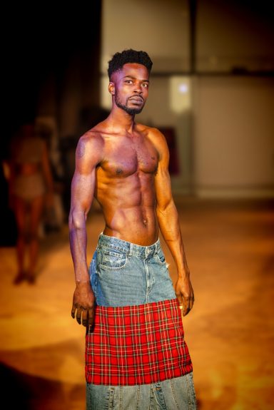 Hombre con torso desnudo, posando con una falda de mezclilla en un entorno artístico.