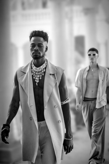 Dos modelos desfilan, uno con collar y abrigo claro, en un entorno elegante en blanco y negro.